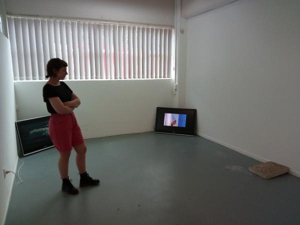 Priscilla Howe, CIRCUIT's 2017 Curatorial Intern, stands in The Audio Foundation gallery looking at an installation of artworks. The works include two videos on separate monitors and a slab of concrete. This is the installation of the exhibition PULSE/REPEAT, a grooup show curated by Priscilla Howe at The Audio Foundation, Auckland (2017).