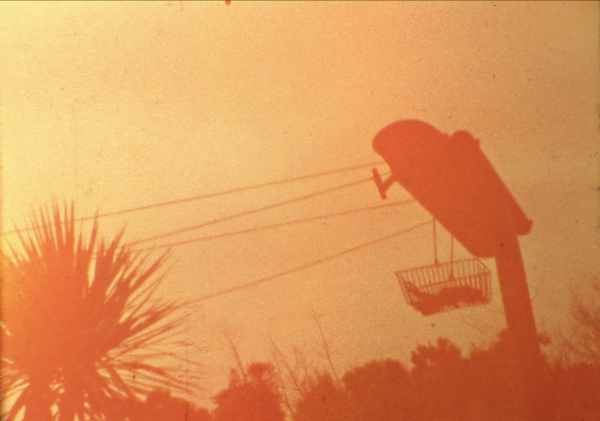 A dark orange shadow of a clothes line appears in contrast against a grainy yellow sky. The clothes line is holding a supermarket shopping basket. In the background there are trees. It feels as if this image was taken in someones backyard.