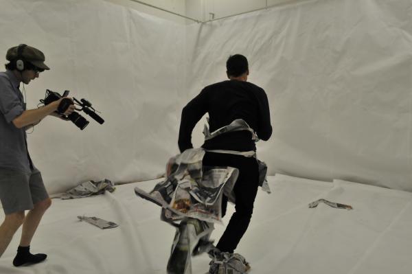 A room lined from floor to ceiling with white paper. At left, a camera operator is shooting a video of the artist Mark Harvey running towards a wall. Mark Harvey is wearing the tattered remains of a costume made entirely of newspaper. The video operator points their camera towards Harvey's feet, which are still covered in newspaper. A room lined from floor to ceiling with white paper. At left, a camera operator is shooting a video of the artist Mark Harvey running towards a wall. Mark Harvey is wearing the tattered remains of a costume made entirely of newspaper. The video operator points their camera towards Harvey's feet, which are still covered in newspaper.
