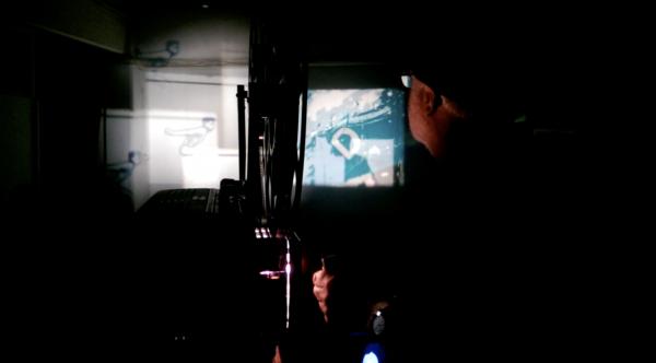 Australian film artist Dirk de Bruyn operates a 16mm film projector in a live performance setting. The projector beams an abstract image onto a wall opposite the projector. We see the projected image in the centre of the picture, between the projector and the operator. The screening is taking place in the dark, the only light is from the projector lamp and projected image. Australian film artist Dirk de Bruyn operates a 16mm film projector in a live performance setting. The projector beams an abstract image onto a wall opposite the projector. We see the projected image in the centre of the picture, between the projector and the operator. The screening is taking place in the dark, the only light is from the projector lamp and projected image.