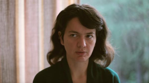 A person with dark hair sits in a domestic setting facing the camera. Their eyes look to something offscreen. The background is blurred, but shows, at left, a pastel coloured curtain. At right, we see a window through whichappears to be nature. A person with dark hair sits in a domestic setting facing the camera. Their eyes look to something offscreen. The background is blurred, but shows, at left, a pastel coloured curtain. At right, we see a window through whichappears to be nature.