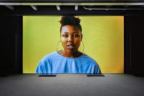 A screen in a darkened shows a Black woman who wears large hoop earrings and a blue top.