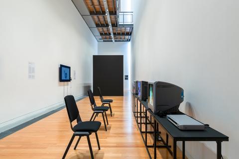 An art gallery with three monitors running along the right-hand wall, one monitor on the left wall, and a black wall at the end.