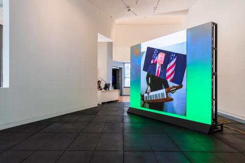 In a gallery space a big screen shows an image of Donald Trump standing in front of the American flag. His torso is a different body and is playing the keytar.