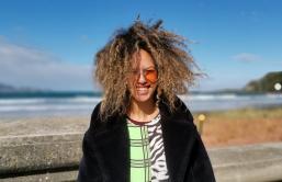 A photo of Israel Randell standing facing the camera. She wears sunglasses and her hair is blown by the wind. Behind her is the ocean.