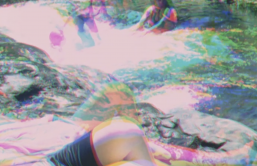 People play near a river or waterfall, laying near the water or swimming. The image has been made with a photographic technique called three colour separation, in which the scene is filmed three times, and each negative given a red, blue and green primary colour. These negatives are layered on top of each other, and combined into one image, which appears to show an ‘after-image’ of different movements recorded in each shot.